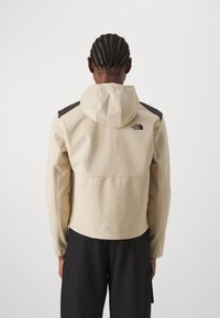 Chaqueta tipo sudadera color beige con una textura suave, acentos negros en los hombros y un logo en la parte posterior. Cuenta con capucha y diseño ajustado.