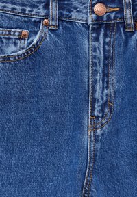 Jean en denim bleu en gros plan montrant la poche avant, les rivets en cuivre, le bouton avec le nom de la marque et les détails de couture orange.