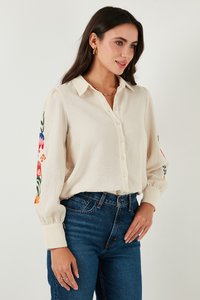 Camisa de botonadura en color crema con mangas largas, decorada con bordados florales en los brazos. Tejido ligero con un acabado texturizado.
