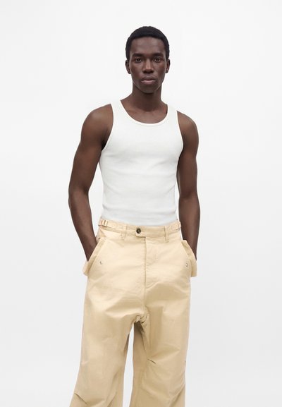 Homme portant un débardeur blanc sans manches et un pantalon beige ample, les mains dans les poches, sur un fond blanc uni.