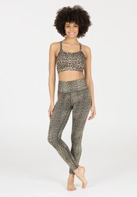 Vrouw staat blootvoets, glimlachend, gekleed in een bijpassende sportbeha en high-waisted leggings met luipaardprint tegen een witte achtergrond.