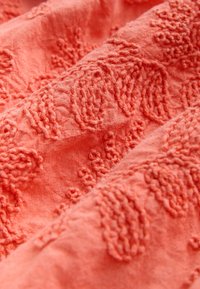 Tissu en corail doux avec broderies florales texturées en relief dans des motifs tourbillonnants créant une surface tactile.