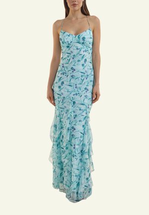 Frau trägt ein hellblaues, bodenlanges Blumenkleid mit dünnen Trägern und geschichteten Rüschendetails vor hellem, einfarbigem Hintergrund.