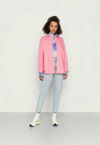Camisa oversized rosa com botões, gola alta em degradê azul, jeans de denim claro e ténis brancos com detalhes em neon, em pé sobre um fundo claro.