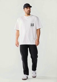Man wearing white oversized T-shirt with black Japanese text, black pants, white sneakers, and black cap standing against plain background.
