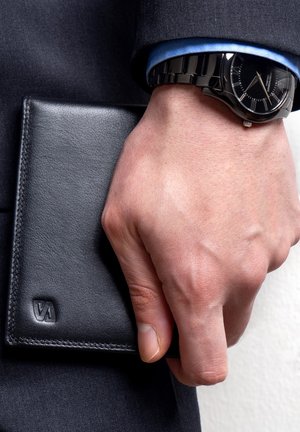 Black leather wallet held in a hand, featuring a subtle embossed logo and stitching detail. A black watch is worn on the wrist.