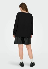 Sieviete stāv, skatoties prom, tērpusies vaļīgā melnā garroku tops, melnas ādas šorti, baltas zeķes un melni masīvi apavi uz baltas fona.