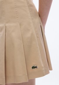 Khaki plooirok gemaakt van een katoenmenging, met een geborduurd groen krokodillenlogo aan de zoom aan de linkerkant.