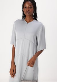 Graues Kurzarmkleid mit hohem Kragen, das subtile Nahtdetails aufweist. Das Model hält eine kleine Handtasche mit metallic-muster.