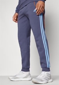 Pantalones deportivos azul marino con rayas laterales de color azul claro, confeccionados en una tela suave. Cuentan con puños elásticos y un cordón naranja en la cintura.