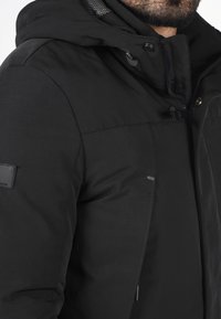 Sort jakke med en struktureret overflade, stor hætte og kontrastfarvet for. Har lynlås, trykknapper og et logo-patch på ærmet.