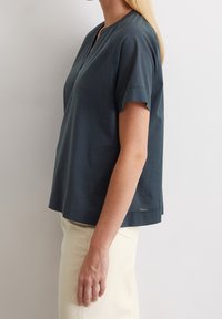 Mörk teal kortärmad topp med v-ringning, avslappnad passform och sidoklipp i fållen, tillverkad av lättviktig tyg. Bärs över en ljusfärgad kjol.