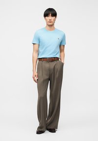 Νεαρός άνδρας που φοράει ανοιχτό γαλάζιο εφαρμοστό μπλουζάκι, καφέ ζώνη, φαρδιά καφέ παντελόνια και σκούρα παπούτσια, στέκεται με το ένα χέρι στην τσέπη μπροστά σε λευκό φόντο.