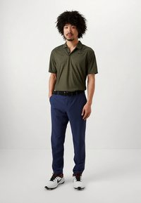 Olivgrön pikétröja med korta ärmar och texturerat mönster, kombinerad med marinblå byxor och vita sportskor med mörka detaljer.