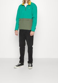 Pullover in pile color teal con collo giallo e parte inferiore verde oliva; presenta una cerniera parziale e tasche frontali, abbinato a jeans neri e sneakers.