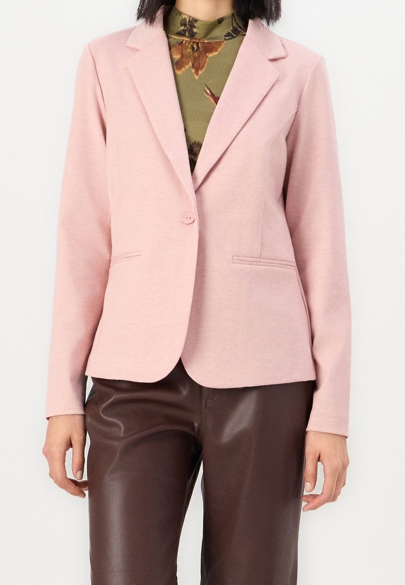 Femme portant un blazer rose clair avec un seul bouton sur un top fleuri vert et un pantalon en cuir marron foncé.