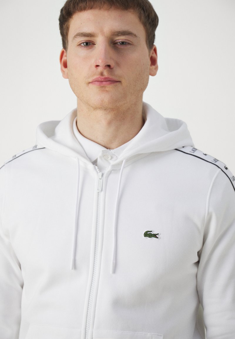 La felpa bianca presenta una chiusura con zip, un cappuccio regolabile con cordino e un logo del coccodrillo verde Lacoste sul petto. I dettagli laterali includono strisce nere.