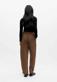 Asmuo su ilgais juodais plaukais, vilkintis juodą ilgomis rankovėmis marškinį, rudus laisvus kelnes ir juodas batus, stovi pasukęs nugarą ir laikydamas rankas kišenėse.