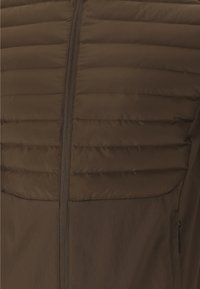 Bruine geïsoleerde jas met een quilted ontwerp, een ritssluiting aan de voorkant en een gladde stof. Het heeft een zijzak met een discrete ritssluiting.