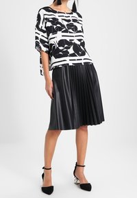 Haut ample noir et blanc avec motif floral et rayé, associé à une jupe plissée en faux cuir noir. Chaussures noires avec brides à la cheville.