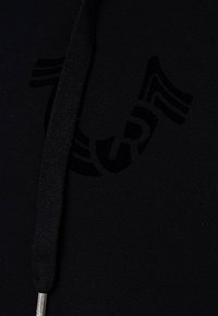 Tissu de sweat à capuche noir avec lettres "SR" en relief et motif rayé, muni d'un cordon de serrage noir avec embout métallique.