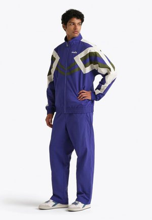 Uomo in piedi che indossa una tuta blu con strisce a chevron bianche e verdi, e sneakers bianche con dettagli blu.