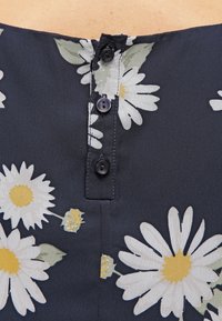 Květinový top vyrobený z tmavě modré látky s bílými kopretinami a žlutými akcenty, s třemi černými knoflíky u výstřihu a hladkou texturou.