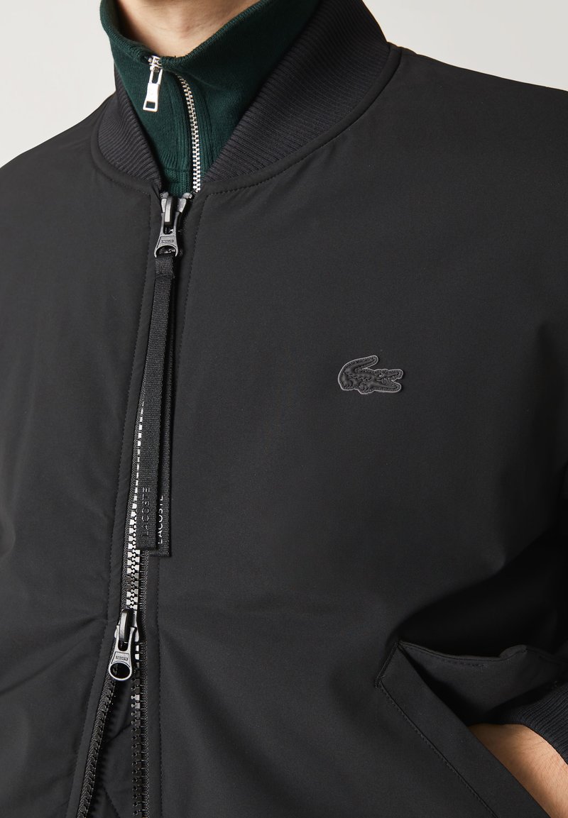 Veste bomber noire en tissu lisse, dotée d'un col montant avec doublure intérieure verte, fermée par un zip à l'avant, et d'un logo en relief sur la poitrine.