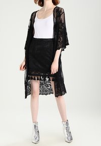 Černý kimono styl svetr s širokými rukávy, s květinovými vzory a třásněmi na spodním lemu, nošený přes černou sukni a bílý top.