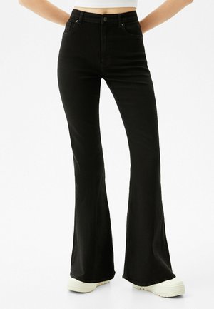 High-waisted black flare jeans worn with white platform sneakers and a cropped white top, shown from waist to feet.