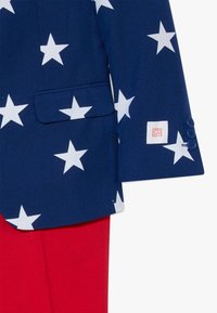 Tumes sinine bleiser valgete tähede mustriga, ühe taskuga ja nööbikaunistustega. All on nähtavad punased püksid. Etikett kannab kirja "OPPO SUITS."