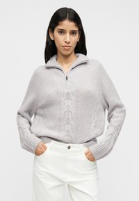 Ανοιχτό γκρι πουλόβερ με πλέξη καλτσοδέτα, ψηλό γιακά και κλείσιμο με φερμουάρ. Μανσέτες και τελείωμα με ριμπ ύφανση. Συνδυάζεται με λευκό παντελόνι. Χωρίς ορατά σχέδια.