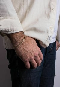 Brățară din aur cu un design răsucit și un charm rotund, purtată pe un încheietură. Mana este sprijinită pe o țesătură deschisă la culoare.