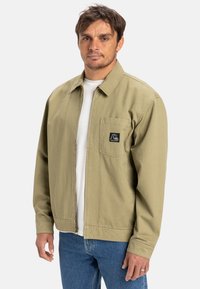 Licht olijfgroene jas met een rits aan de voorkant, borstzak en een klein logo-embleem. Gemaakt van een duurzame stof met een relaxte pasvorm.