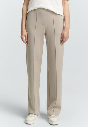 Beige weite Hosen mit Seitenfalten und einer glatten Textur, ausgestattet mit einer taillierten Taille und dezenten Nähdetails, kombiniert mit weißen Sneakers.