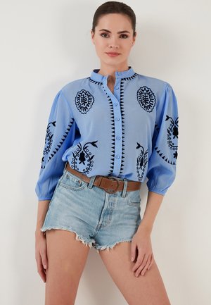 Blue button-up shirt with black floral embroidery and puffed sleeves, paired with distressed denim shorts and a brown belt.
