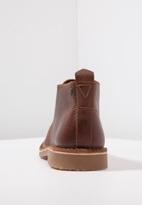 Hnědé kožené kotníkové boty s texturovaným povrchem, kulatou špičkou, kontrastním šitím a kovovým detailem na zadním podpatku. Plochá gumová podrážka.