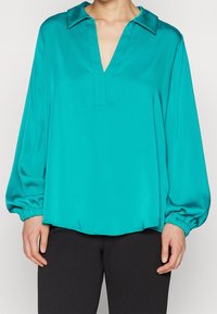 Teal satin blouse with a V-neck, oversized sleeves, and elastic cuffs, featuring a smooth texture and casual fit, worn over black trousers.