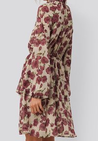 Robe florale avec de longues manches bouffantes, en tissu beige crémeux orné de roses bordeaux, une taille cintrée et une jupe superposée.