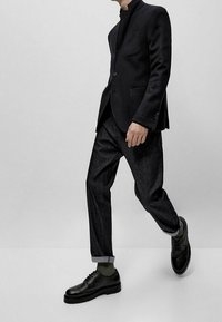 Svart skräddarsydd kavaj med hög krage, kombinerad med mörka denimbyxor och svarta snörskor. Strukturerad tyg och uppvikta ärmar synliga.