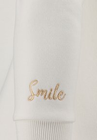 Tessuto bianco a coste con un polsino che presenta un testo ricamato in oro che dice "Sorridi." La texture è liscia con un leggero strech.