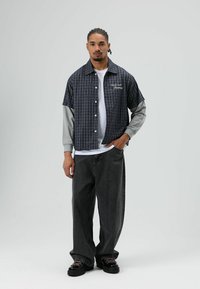 Korte mouwen geruit overhemd in marineblauw en grijs met knopen, gedragen over een wit shirt met lange mouwen, gecombineerd met donkergrijze broek met wijde pijpen.