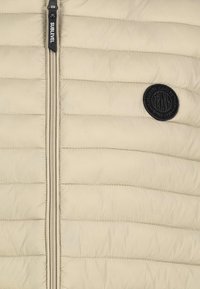 Beiger, gestepptes Material mit horizontalen Nähten, schwarzem, runden Markenpatch auf der rechten Seite und vertikalem Reißverschluss mit "Sublevel"-Etikett auf der linken Seite.