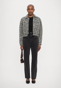 Melnā un baltā houndstooth raksta īsā jaka, pieguļošs melns tops, augstiem jostas vietām tumsas džinsi, melnas plakanas kurpes un neliela brūna rokas soma.