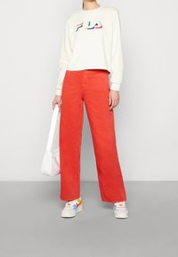 Weißes Sweatshirt mit buntem FILA-Logo, kombiniert mit weit geschnittenen orangen Hosen. Das Modell hält eine weiße Tasche und trägt mehrfarbige Sneakers.