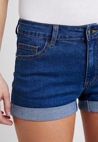 Pantalones cortos de denim en azul oscuro con dobladillo enrollado. Incluye un bolsillo delantero, pasadores para cinturón y costuras en contraste. Textura suave.