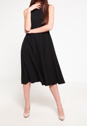 Czarna sukienka bez rękawów o zwiewnym, rozkloszowanym kroju w kształcie litery A. Wykonana z lekkiego materiału, z dołem sięgającym poniżej kolan, zestawiona z beżowymi szpilkami.