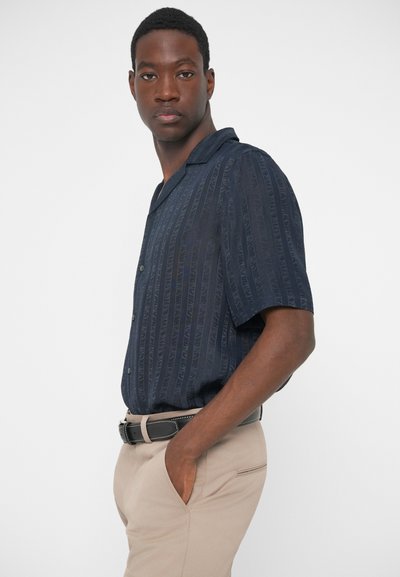 Μπλε πουκάμισο κοντομάνικο με κατακόρυφες ρίγες, άνετη εφαρμογή, μπροστινός κουμπωτός κλειστός λαιμός, μαλακή υφή. Φορεμένο με μπεζ παντελόνι και μαύρη ζώνη.