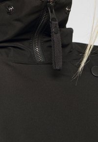 Giacca nera con colletto alto, zip frontale e dettagli con bottoni a pressione. Presenta una texture liscia e opaca e un cordino regolabile.