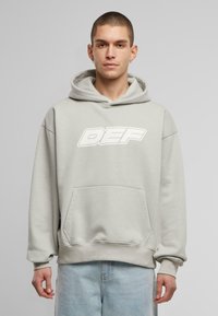 Lys grå hoodie med en frontlomme og et stort hvidt "DEF" grafik på brystet. Har lange ærmer og en rummelig hætte.